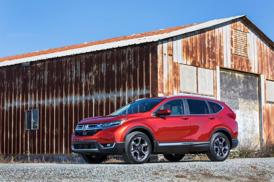 A metallic red 2018 Honda CR-V parked on gravel in front of a rusty barn and blue sky