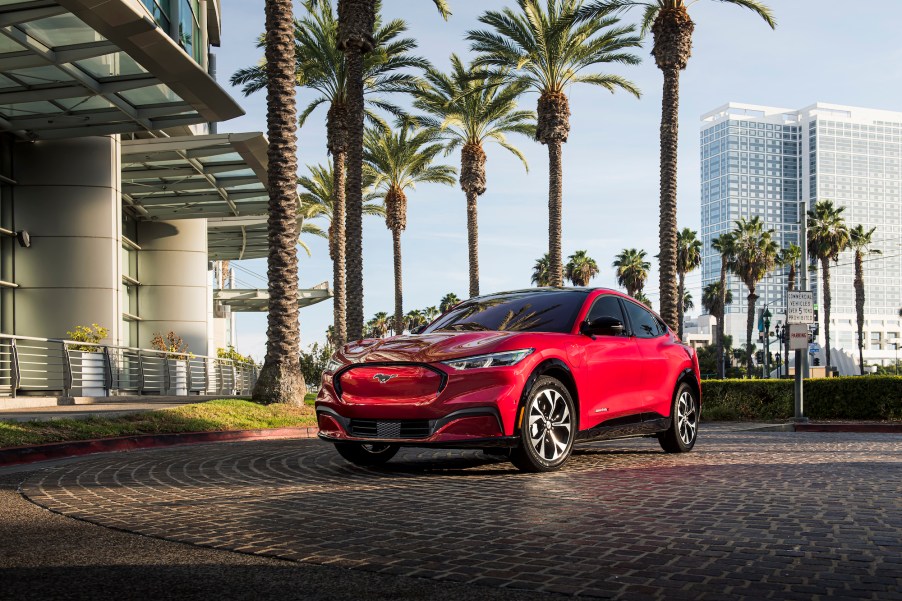 A red 2021 Ford Mustang Mach-E parked in a circular driveway outside a modern building lined with palm trees