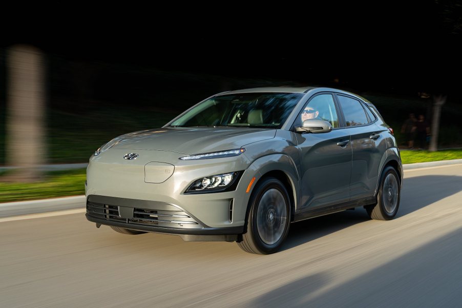 A gray 2022 Hyundai Kona Electric compact crossover SUV traveling on a road along manicured grass and trees