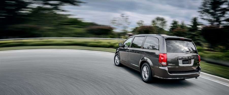 A dark-gray 2020 Dodge Grand Caravan minivan traveling on a road lined with green grass and trees