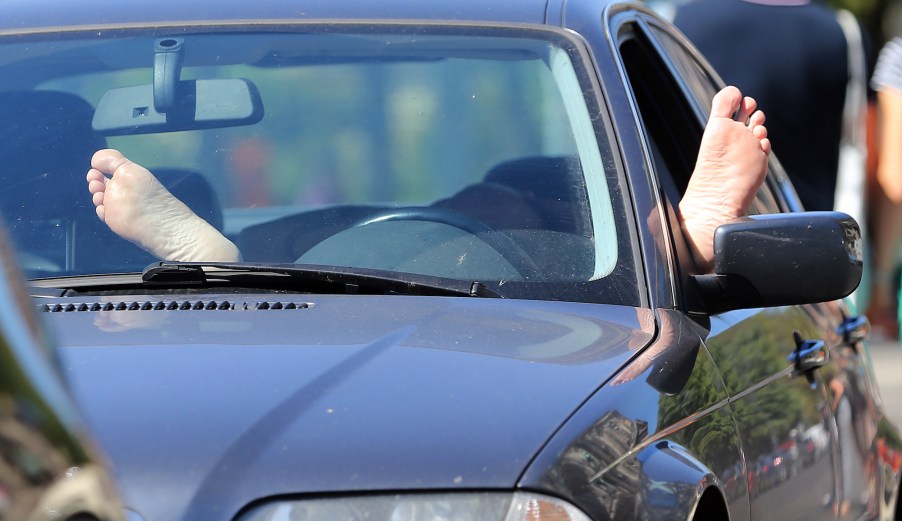 A car driver relaxes behind the steering wheel with bare feet propped up on the dashboard and window sill