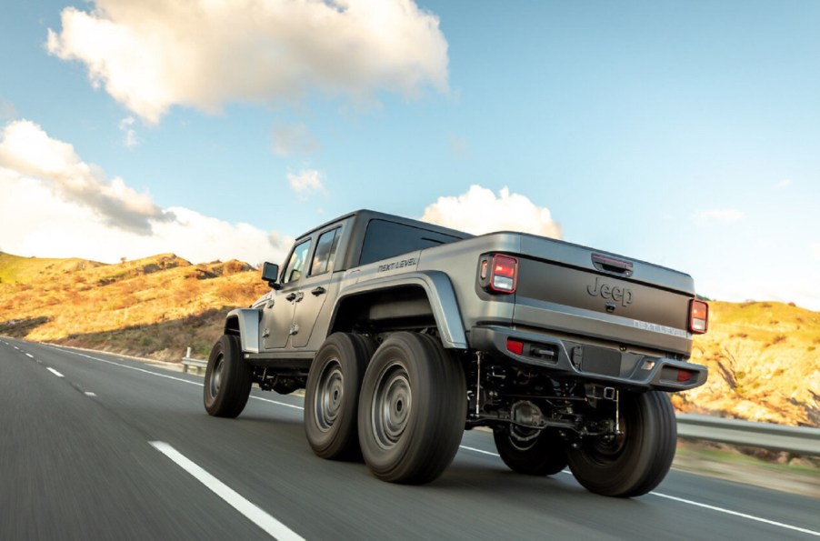 A low-angle rear 3/4 view of a matte-gray Next Level Jeep Gladiator 6x6 driving on a hilly road