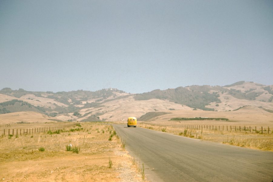 A yellow Volkswagen bus travels along a country road that cuts through a prairie covered in dry grass, low rolling hills can be seen in the background on a summer road trip