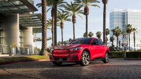 A red 2021 Ford Mustang Mach-E sits on a brick driveway surrounded by palm trees and modern buildings.