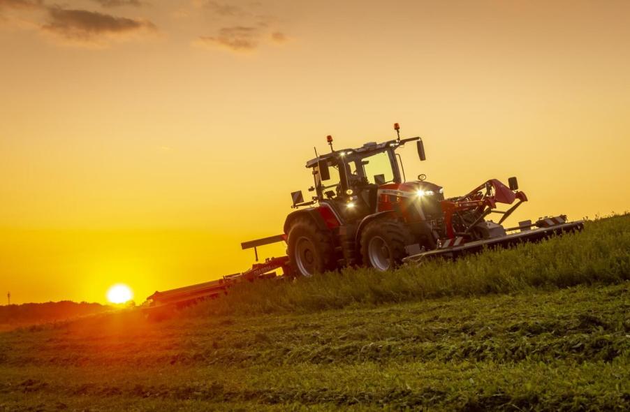 A Massey Ferguson 8S tractor working a hay field at sunrise