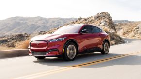 A red 2021 Ford Mustang Mach-E is driving on a highway with a brown rocky scene in the background.