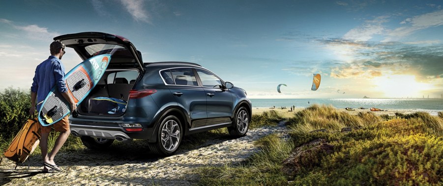 A blue 2022 Kia Sportage parked in front of the ocean. A man loads a surfboard into the back of the car.