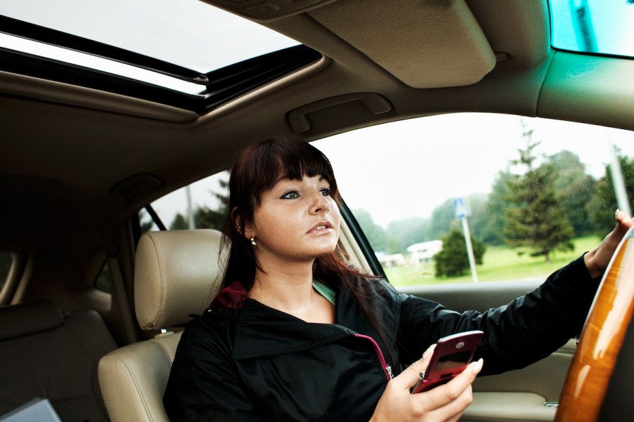 A young woman sits at the wheel of a car while holding a cell phone