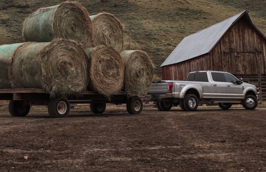 Silver Ford F-Series F 250 Super Duty truck towing a trailer loaded with hay bales in front of a barn.