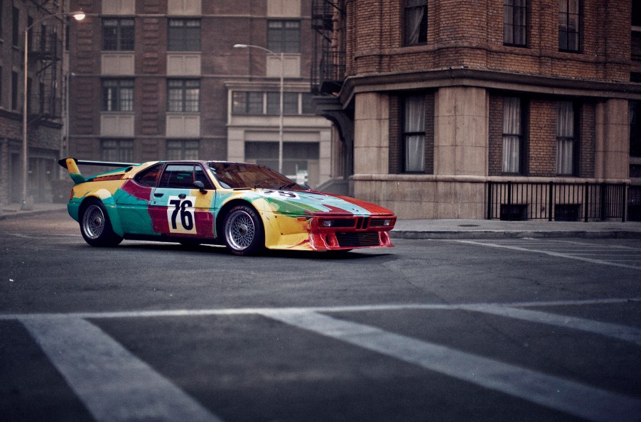 A 3/4 profile view of the Andy Warhol painted 1979 BMW m1 race car. Shown parked on a street with stone buildings in the background.