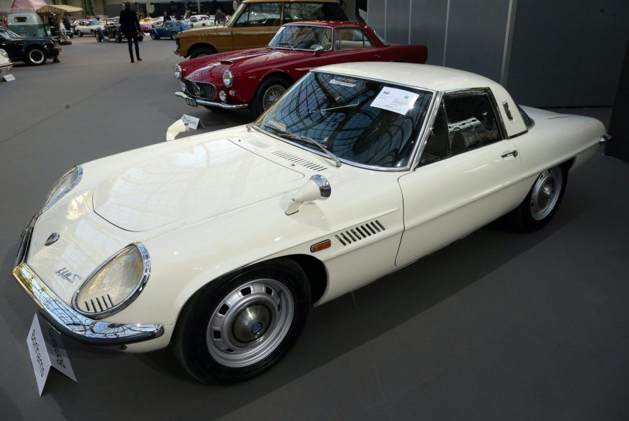 A 3/4 front/profile view of a white 1970 Mazda Cosmo 110S sports car. The car is parked in line with a red sports car at the Grand Palais in Paris, France.