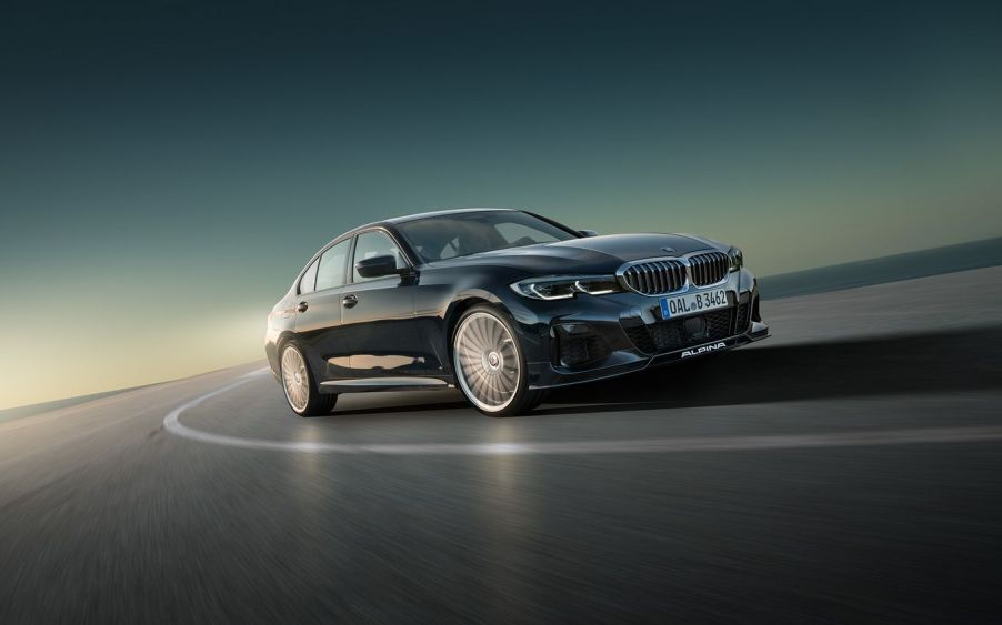 A 3/4 front view of a dark blue Alpina B3 sedan cornering hard on a road.