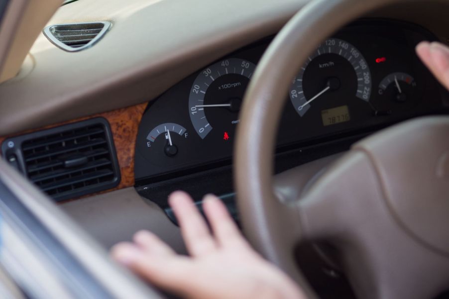 Close up of a half-empty fuel gauge in an older-model-year vehicle; a pair of hands making an exasperated gesture are blurred in the forefront