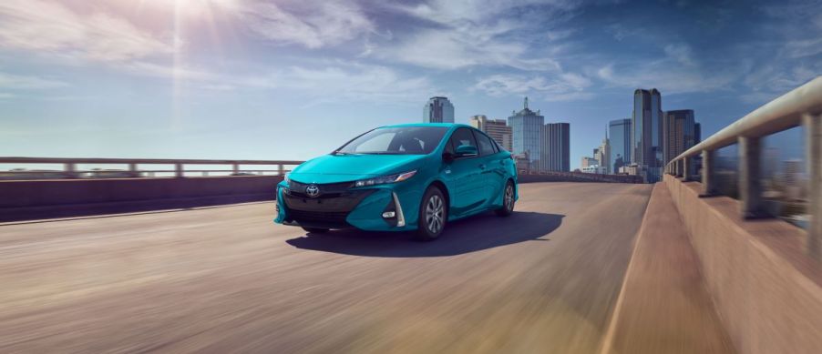 A blue 2022 Toyota Prius Prime driving down a road with a city in the background.