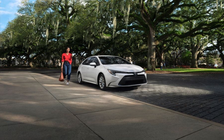 Woman in red holding a coffee walking toward a new white 2023 Toyota Corolla sedan parked on a street