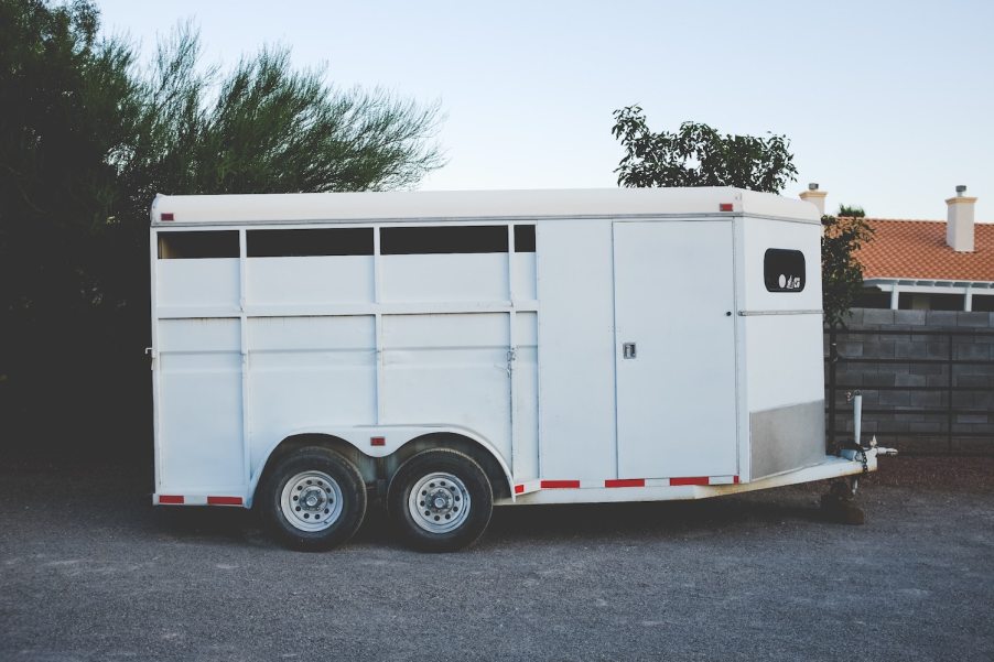 White, tandem axle livestock trailer, parked in front of trees.