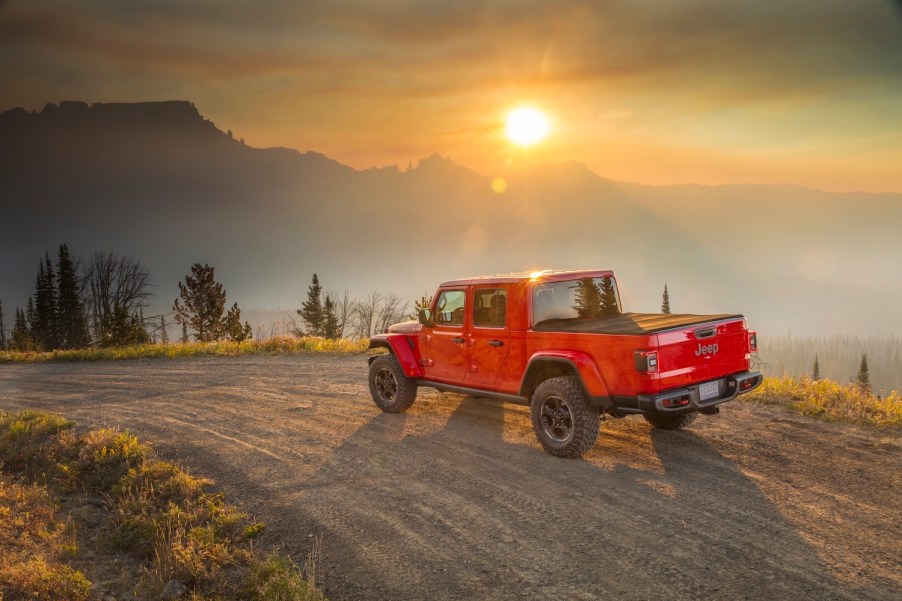 Promo photo of a red Jeep Gladiator 4WD truck navigating a dirt road, the sun setting behind a mountain range visible in the background.