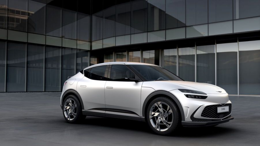 A white Genesis GV60 electric compact SUV model parked on the plaza outside of a glass office building