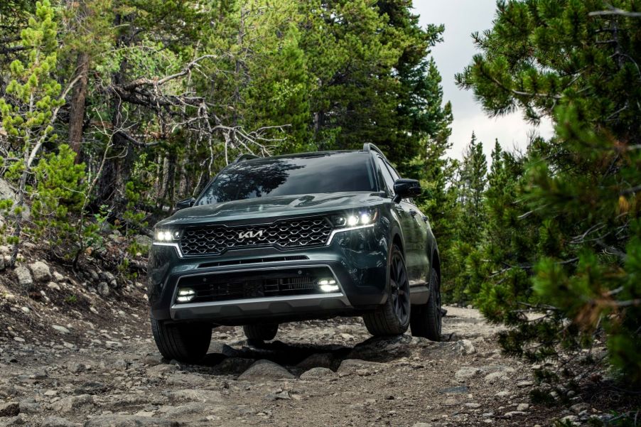 A dark green 2023 Kia Sorento X-Line compact SUV model driving on a gravel dirt road in a forest