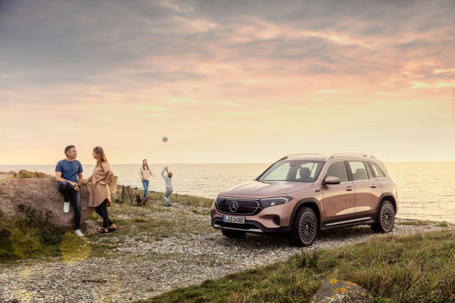 A bronze/copper Mercedes-Benz EQB luxury SUV model parked on a gravel road near a sunset beach as a family plays