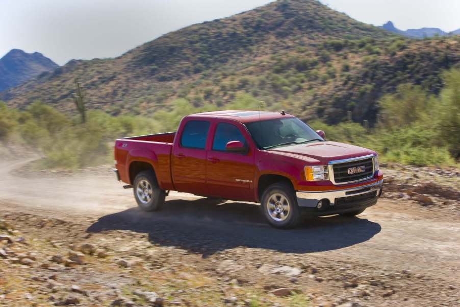 A red GMC Sierra 1500 pickup truck driving up a dirt road, mountains visible in the background.