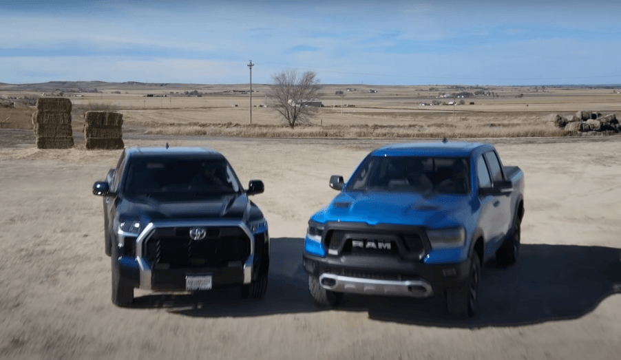 A dark blue Toyota Tundra and light blue Ram 1500 parked next to one another on a farm, hay bales in the background.