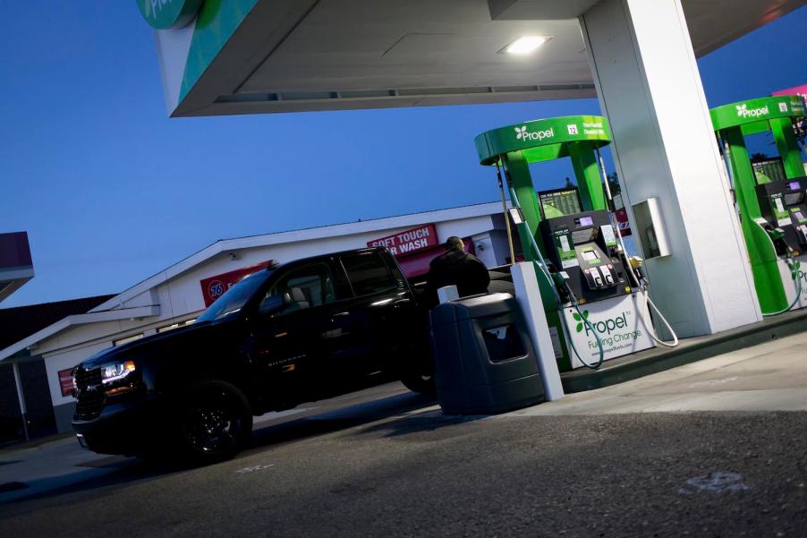 Man gases up his black Silverado pickup truck at a station that sells diesel, the sunset visible in the background.