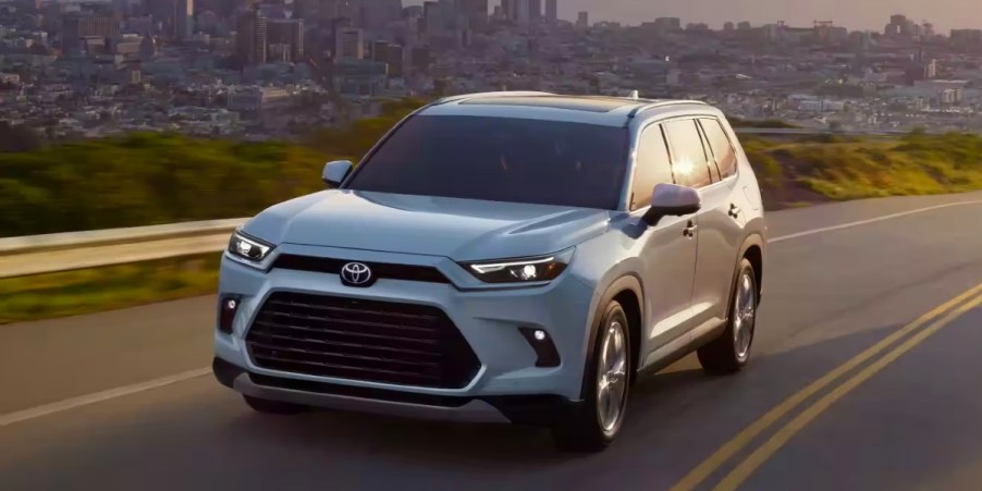 A white 2024 Toyota Grand Highlander midsize three-row SUV is driving on the road.