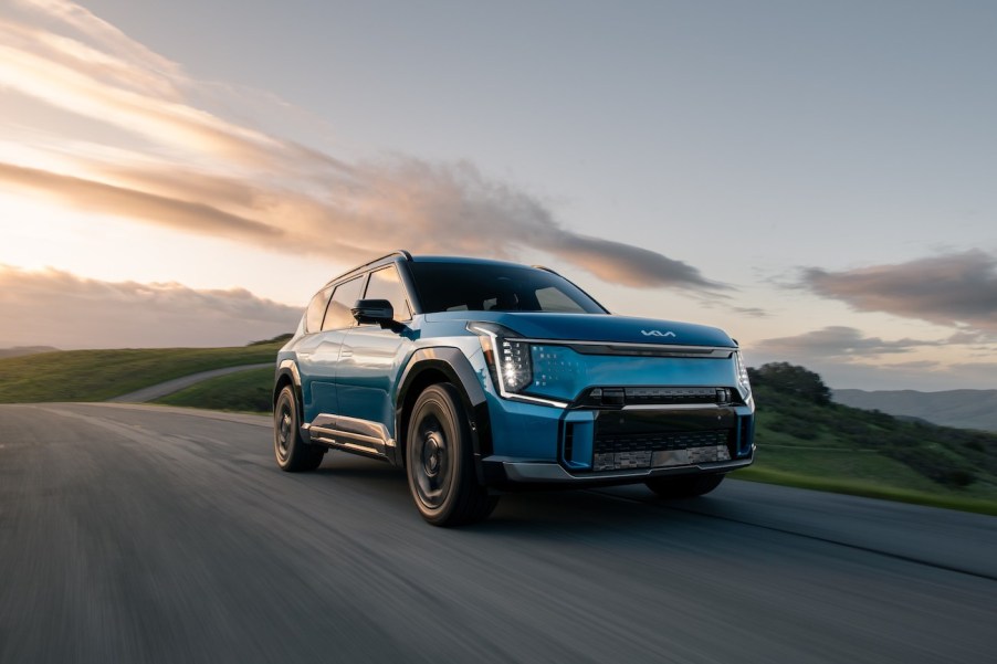 A blue Kia EV9 electric SUV driving while the sun sets behind it.