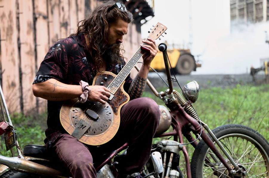 Jason Momoa plays a guitar on a motorcycle.