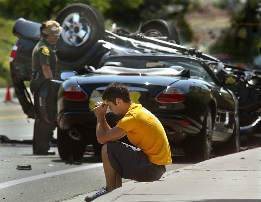 A motorist sits next to a Jaguar X100 XK and a totaled car after a crash.