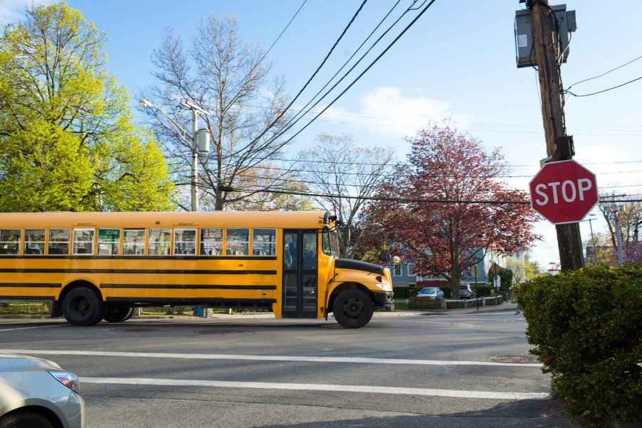 A school bus in nearly full right side profile view entering an intersection with a stop sign posted in the right midground and car nose peeking out of left rear corner