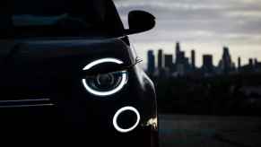 A 2024 Fiat 500e photographed outside in shadow with left headlamp illuminated in close left front side view with cityscape in right frame background