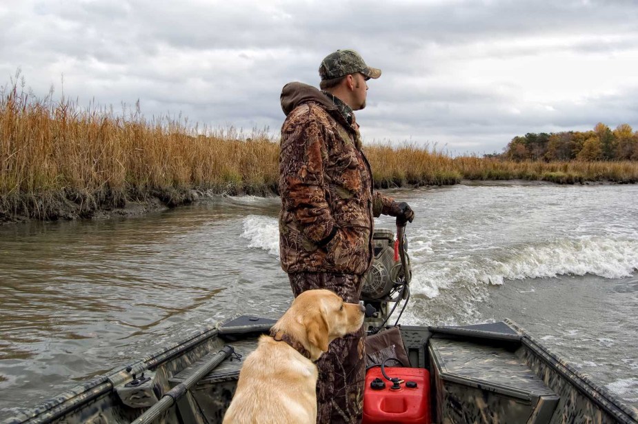 Hunter in camouflage clothes holding the tiller of a powerboat outboard engine while motoring down a river, a dog sitting at his feet.