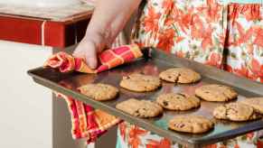 A 1950s-style close up of a woman holding a tray of chocolate chip cookies wearing a flowery apron