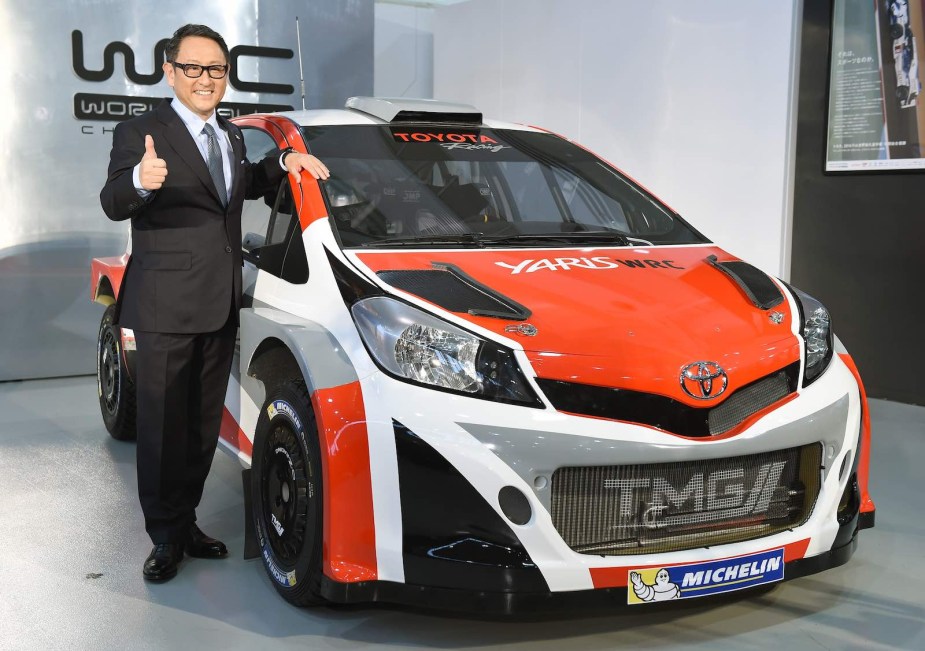 Businessman Akio Toyoda gives the thumbs up standing next to a bright orange Yaris race car.