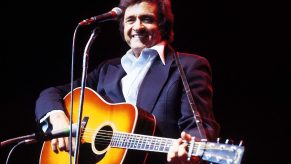 Johnny Cash smiles onstage holding guitar with black background in 1979 wearing white button up and dark jacket