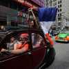 Young woman stands up in the back of a Citroën 2CV driving through Times Square, holding a French flag