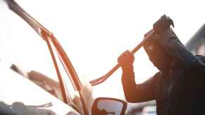 Man dressed in all black with black mask holding a crow bar above the right front window of a car