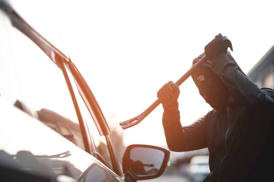 Man dressed in all black with black mask holding a crow bar above the right front window of a car