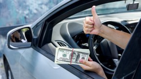 A depiction of a vehicle diminished value claim with a woman's hands holding a stack of money in left hand with right hand doing 'thumbs up' motion out of an open car window