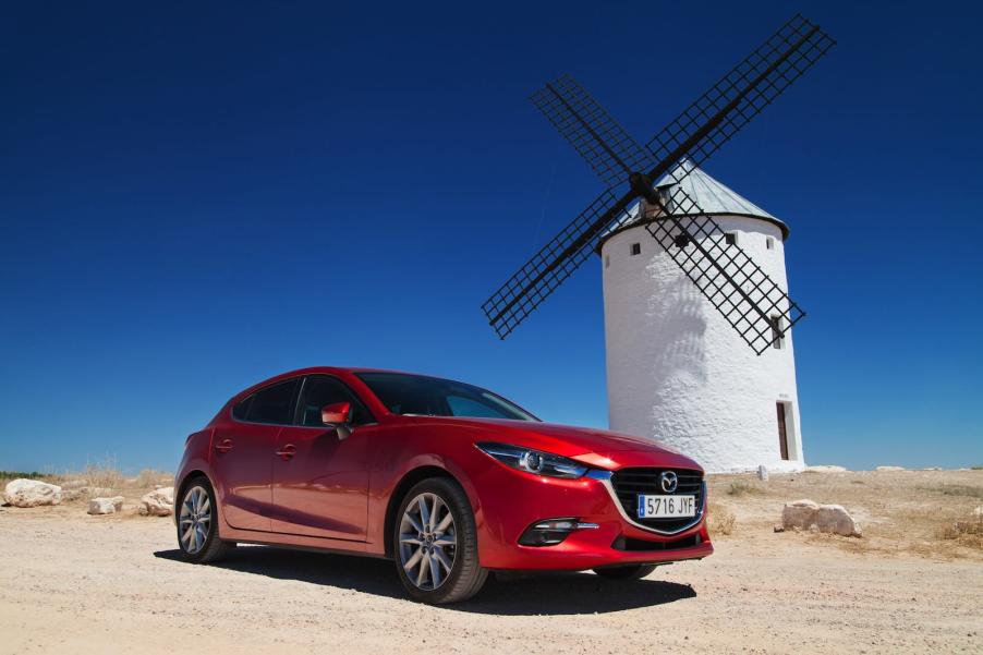 Bright red Mazda3 wagon parked in front of a windmill, the Spanish countryside visible behind it.