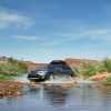 A Subaru Outback drives through a stream, mountains visible in the background.