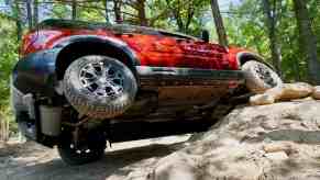 Red 2025 Bronco Sport Badlands shows off its 29-inch Goodyear tires by lifting one in the air as it crawls over rocks, trees visible in the background.