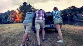 Group of guys push a broken down pickup ruck through a field, trees visible in the background.