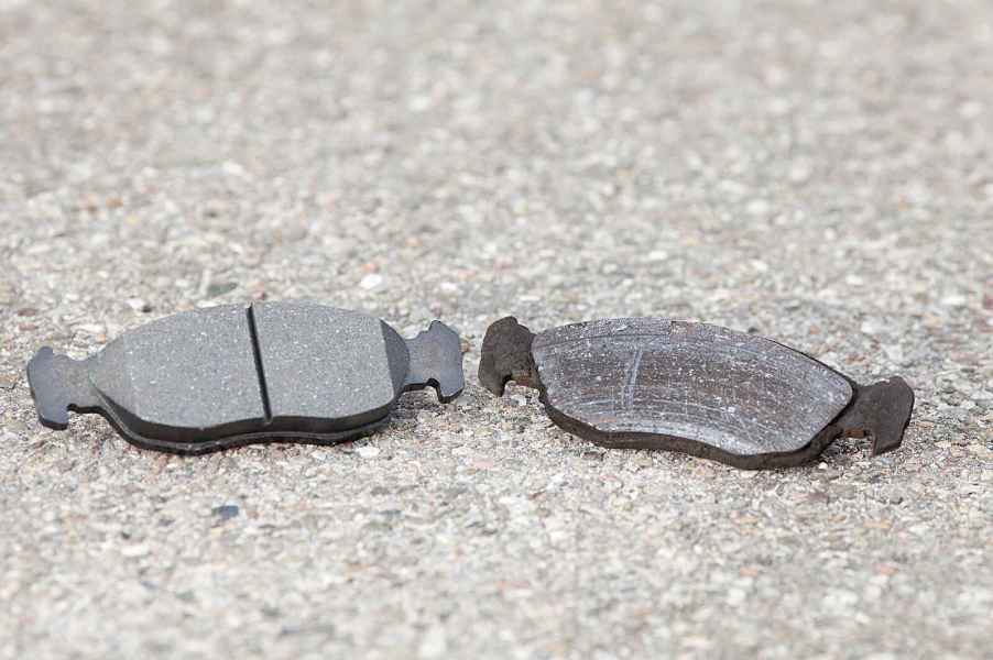 Two car brake pads, one new and one old, placed side by side on pavement in close view