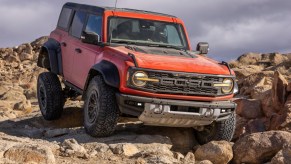 A reddish-orange 2022 Ford Bronco Raptor SUV climbing dusty boulders off-road in right front angle view