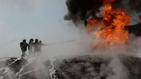 Three firefighters direct a hose at a burning truck, a ball of orange flame above it.