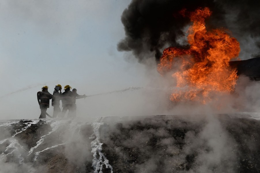 Three firefighters direct a hose at a burning truck, a ball of orange flame above it.