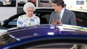 The Queen of England looks at a Jaguar car in surprise while the man next to her talks.